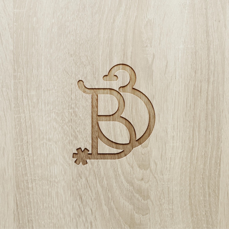 May include: A wooden monogram featuring the letter "B" with a decorative flourish and a star-like element. The design is carved into a light-colored wood surface, creating a subtle contrast in tones.