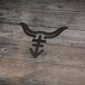 May include: A dark brown graphic of a bull's head with a stylized symbol below it, stenciled on a wooden surface. The wood grain is visible, with a mix of light and dark brown tones.