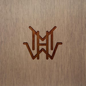 May include: A close-up of a wooden surface with a carved monogram design. The monogram features the letters "M", "H", and "W" in a stylized, symmetrical arrangement. The wood has a light brown color with visible grain.