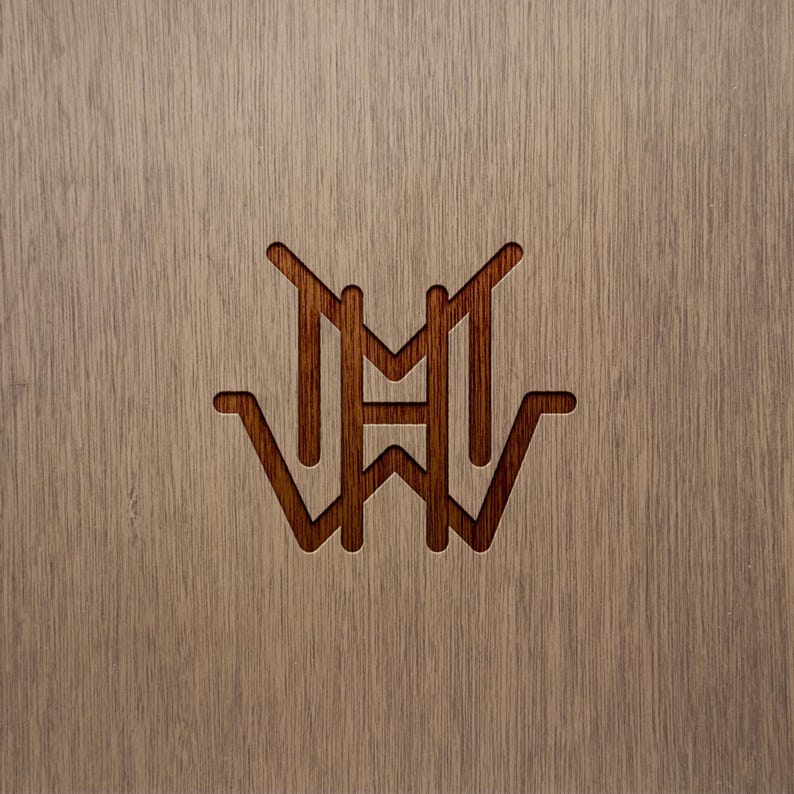 May include: A close-up of a wooden surface with a carved monogram design. The monogram features the letters "M", "H", and "W" in a stylized, symmetrical arrangement. The wood has a light brown color with visible grain.