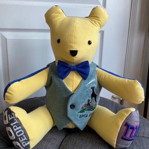 May include: A yellow teddy bear wearing a blue bow tie and a blue and gray vest. The vest has an embroidered image of a dog and the text "SPCA 2017". The bear's feet are made of fabric with the words "PEOPLE" and "M" embroidered on them.