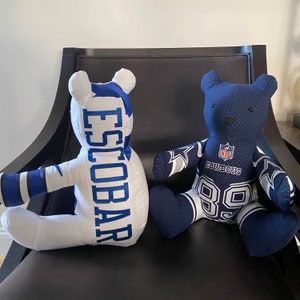 May include: Two plush teddy bears wearing Dallas Cowboys jerseys. One bear is white with the name "ESCOBAR" on the back. The other bear is blue with the number "89" on the front and the NFL logo and "COWBOYS" on the chest.