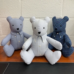 May include: Three teddy bears wearing shirts made from repurposed fabric. The bears are sitting on a wooden surface. The shirts are patterned with stripes and plaid.