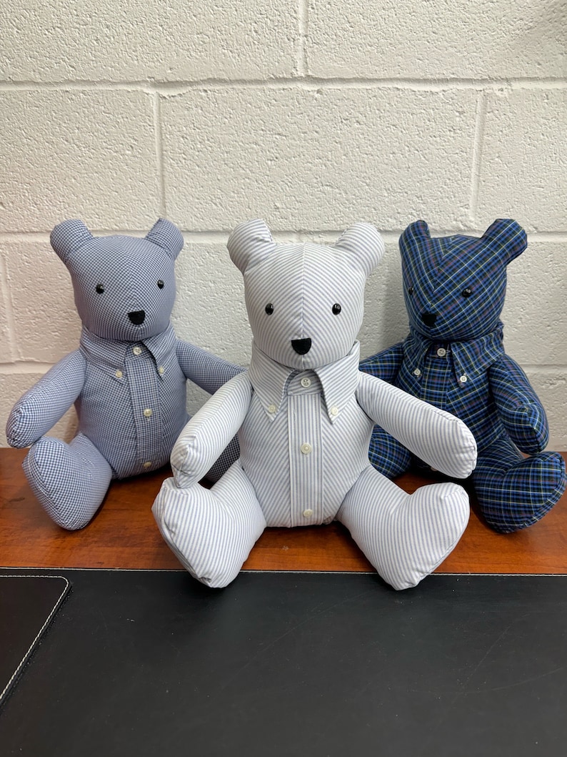 May include: Three teddy bears wearing shirts made from repurposed fabric. The bears are sitting on a wooden surface. The shirts are patterned with stripes and plaid.