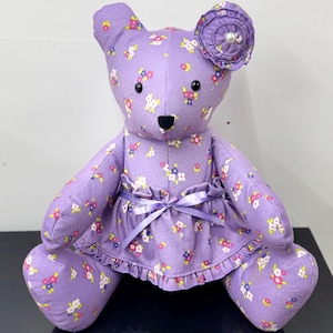 May include: A handmade purple teddy bear with a floral pattern. The bear wears a matching dress with a ruffled hem and a satin ribbon bow. A fabric flower with a pearl center adorns one ear. The bear is approximately 12 inches tall.