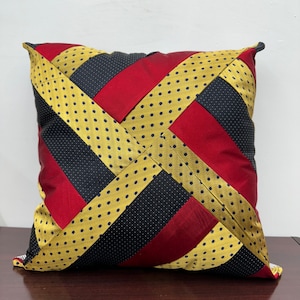 May include: A decorative pillow with a geometric pattern made from repurposed neckties. The pillow is made of black, red, and gold fabric with a polka dot pattern.