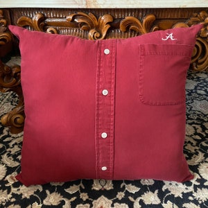 May include: A red pillow with a button-down shirt design and a pocket. The pillow has white buttons and the letter "A" embroidered in white on the top right corner.