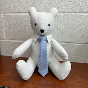 May include: A white stuffed teddy bear wearing a white shirt and a blue patterned tie. The bear is sitting on a wooden surface.