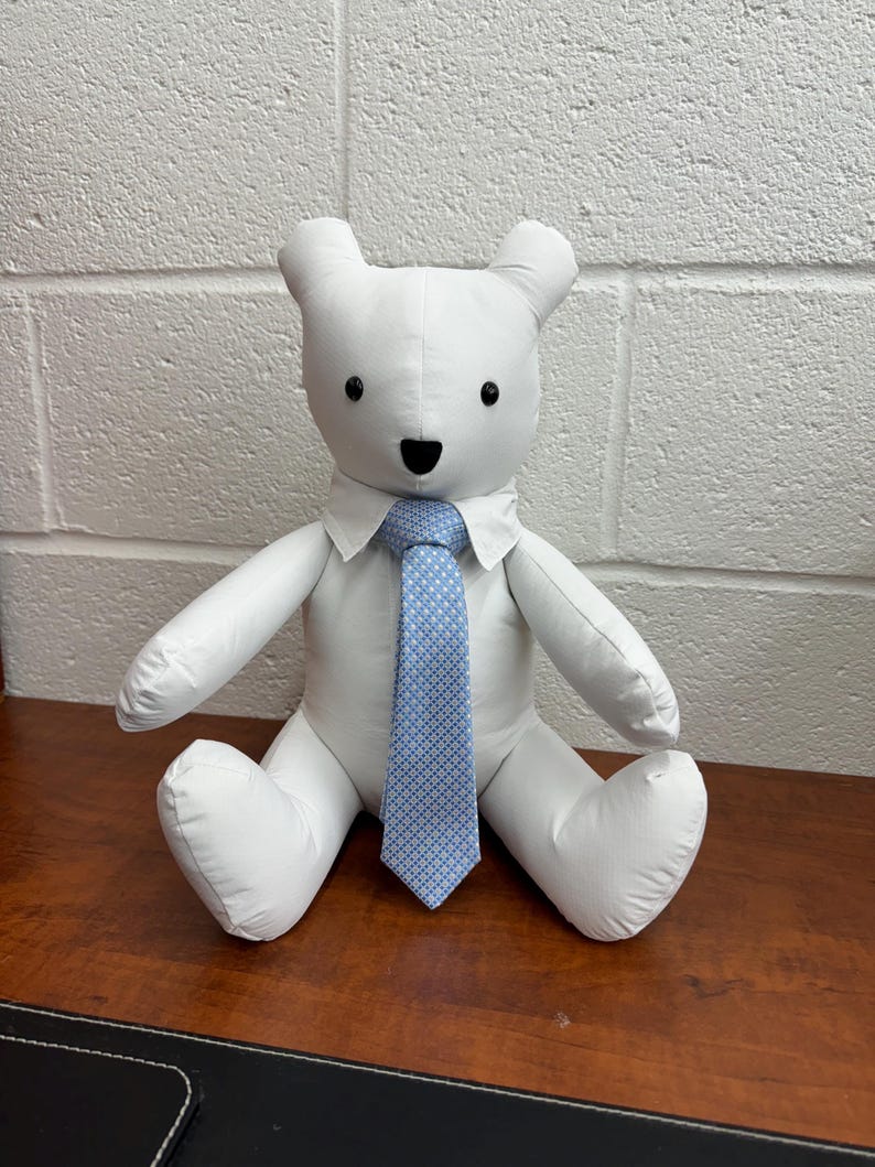 May include: A white stuffed teddy bear wearing a white shirt and a blue patterned tie. The bear is sitting on a wooden surface.