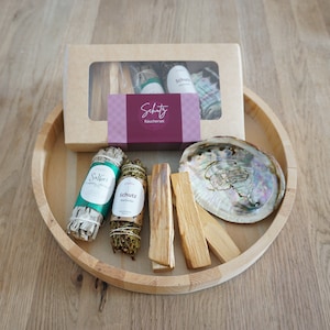 Peut inclure: Un plateau en bois contenant une variété de bâtonnets de sauge, dont de la sauge, du cèdre et du palo santo. Il y a également une coquille et une boîte d'encens étiquetée "Schutz Räucherset".
