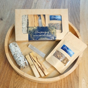Könnte beinhalten: Ein Holztablett mit einem Hausreinigung-Set mit Salbeibündeln, Palo Santo-Stäben und kleinen Kräutertüten. Eine Pappschachtel mit Fenster zeigt ähnliche Artikel. Ein klarer Kristallstab ist enthalten.