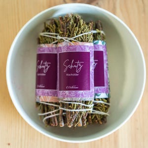 Könnte beinhalten: Drei Bündel Wacholder-Reinigungsstäbchen, umwickelt mit weißem Garn und mit lila und weißen floralen Musterpapier beschriftet. Die Etiketten lauten "Schutz Wacholder" und "WellDone".