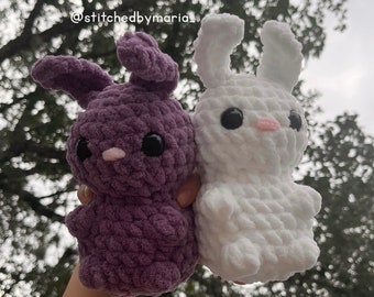 Könnte beinhalten: Zwei gehäkelte Hasen-Plüschtiere, eines lila und eines weiß. Der lila Hase hat schwarze Augen und eine rosa Nase. Der weiße Hase hat schwarze Augen und eine rosa Nase. Die Hasen werden in der Hand einer Person gehalten.