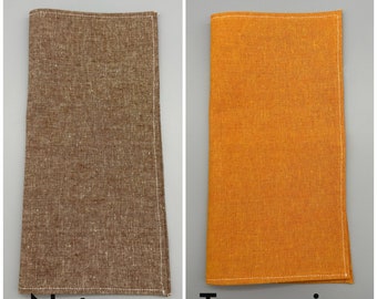 May include: Two folded linen napkins, one in a nutmeg brown and the other in tangerine orange. Both have white stitching along the edges. The text "Nutmeg" and "Tangerine" are printed below each napkin.