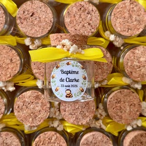 Può includere: Piccoli vasetti di vetro con tappi di sughero, decorati con nastri gialli e un'etichetta con la scritta "Baptême de Clarke 23 août 2026" con illustrazioni di api. I vasetti sono riempiti con una sostanza sconosciuta.