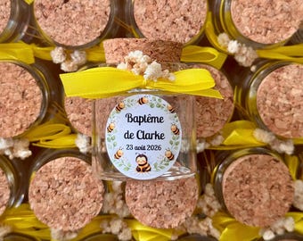 May include: A small glass jar with a cork stopper, featuring a yellow ribbon and a white oval label. The label displays a wreath of green leaves, small bee illustrations, and the text "Baptême de Clarke 23 août 2026" in black font.