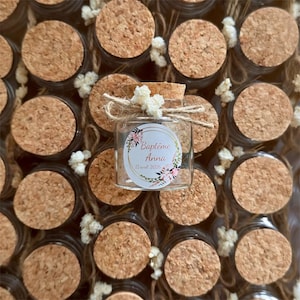 Peut inclure: Petits pots en verre avec couvercles en liège, disposés de près. Un pot est mis en évidence, orné d'une étiquette indiquant "Baptême Anna 12 mars 2026" dans une couronne florale. Le pot est noué avec de la ficelle et de petites fleurs blanches.