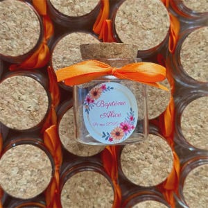 Peut inclure: Un bocal en verre transparent avec un bouchon en liège et un ruban orange noué. Le bocal porte une étiquette circulaire avec une couronne florale rose, orange et bleue, affichant le texte Baptême Alice et la date 24 mai 2026.