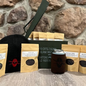 Op de afbeelding: Een groene metalen munitiebox met de deksel open, gevuld met kleine bruine papieren zakjes met verschillende smaken beef jerky. Er is een zwarte beanie, een bruine koozie met een schedel en gekruiste beenderen logo, en verschillende zakjes beef jerky op een houten oppervlak.
