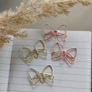 Op de afbeelding: Vier gouden en roségouden strikvormige paperclips op een wit, gestreept notitieboekje. De clips zijn decoratief en kunnen worden gebruikt om papier bij elkaar te houden.