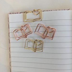 Op de afbeelding: Vier metalen paperclips in de vorm van een boek met hartaccenten. Twee zijn goudkleurig en twee zijn roségoudkleurig. De paperclips zijn gerangschikt op een gelijnde notitieboekpagina, wat wijst op een thema van briefpapier of kantoorbenodigdheden.
