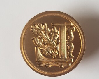 Op de afbeelding: Een gouden metalen waszegelstempel met een sierlijk ontwerp met een gestileerde letter L en bloemmotieven.