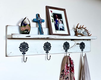 May include: White painted wooden shelf with four black metal hooks and a decorative design. The shelf has a framed photo, a cross, and a glass sculpture on top.