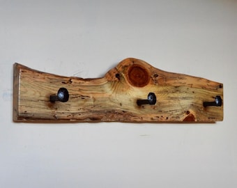 May include: A rustic wooden coat rack with three black metal hooks. The wood has a natural, weathered finish and a unique grain pattern.