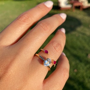May include: A gold ring with a large oval-shaped diamond and two smaller ruby stones on the band. The ring is worn on a person's finger.