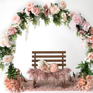 May include: A newborn baby sleeping on a pink faux fur bench, surrounded by a floral archway made of pink and white flowers.