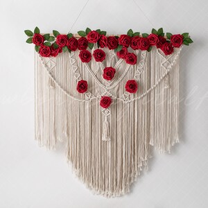 May include: A white macrame wall hanging with a wooden dowel at the top. The dowel is decorated with red silk roses and green leaves. The macrame is in the shape of a heart and has a fringe at the bottom.
