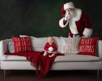 May include: A festive scene with a baby and Santa Claus. The baby sits on a white couch with red pillows that say "FA LA LA" and "MERRY CHRISTMAS." Santa gestures for silence. A red blanket and a pillow that says "HELLO WINTER" complete the holiday setting.