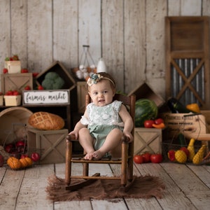 May include: A baby girl wearing a green and white dress sits in a wooden rocking chair. The chair is surrounded by a variety of fruits and vegetables, including tomatoes, watermelon, and zucchini. A sign above the chair reads "farmers market".