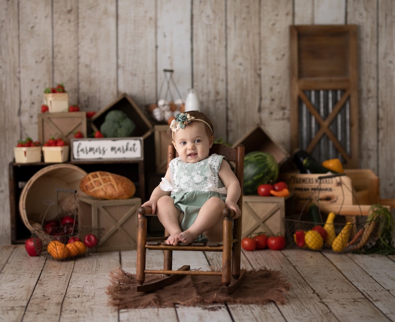 May include: A baby girl wearing a green and white dress sits in a wooden rocking chair. The chair is surrounded by a variety of fruits and vegetables, including tomatoes, watermelon, and zucchini. A sign above the chair reads "farmers market".