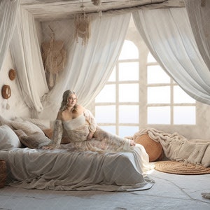 May include: A woman in a white dress sits on a bed with white linens and a white canopy. The bed is in a room with white walls and a large window with white curtains. The room is decorated with macrame wall hangings and other bohemian decor.