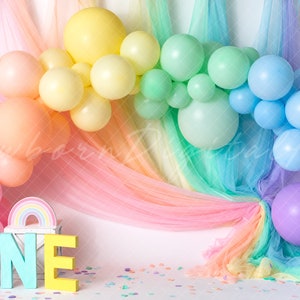 May include: A colorful birthday party backdrop featuring a balloon arch in pastel shades of pink, yellow, green, and blue. The word "ONE" is displayed in large, colorful letters, with a rainbow accent. The background includes rainbow-colored fabric.