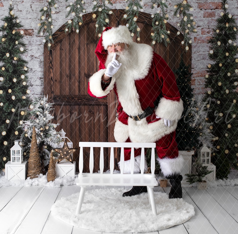 May include: Santa Claus in a red suit with white trim and a white beard is standing next to a white wooden bench. He is holding his finger to his lips as if to say 'shhh'. The background is a brick wall with a wooden door and Christmas trees.