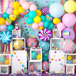 May include: A colorful backdrop with a rainbow, balloons, and candy-themed decorations. There are white boxes with candy and other decorations on shelves. The backdrop is perfect for a birthday party or a candy-themed event.