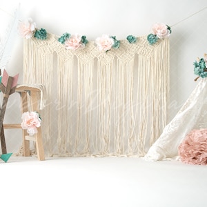 Op de afbeelding: Een witte achtergrond met een macramé wanddecoratie versierd met roze en groene bloemen. Twee houten krukjes met roze bloemen en houten pijlen staan aan de linkerkant van de afbeelding. Een witte tipi met roze tissuepapierballen staat aan de rechterkant van de afbeelding.