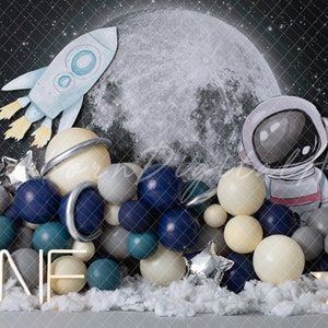 May include: A space-themed first birthday party backdrop featuring a large moon, a rocket, an astronaut, and a balloon arch in shades of blue, grey, and white. The balloon arch includes a silver star and a white sign that says "ONE".