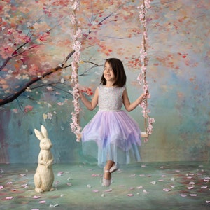 May include: A young girl in a lavender and blue tulle dress sits on a swing in front of a painted backdrop of a blooming cherry blossom tree. A white rabbit figurine sits on the ground in front of her.