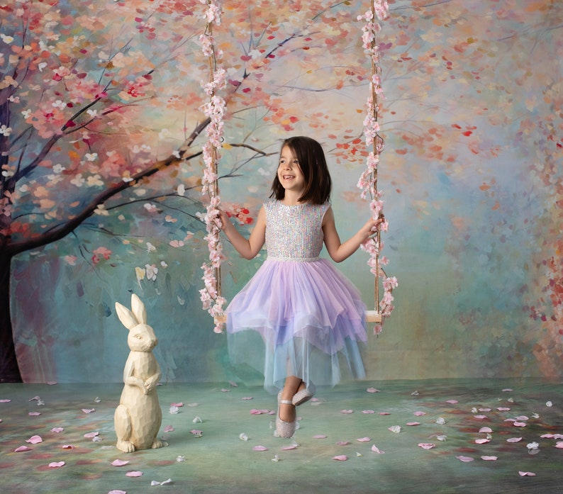 May include: A young girl in a lavender and blue tulle dress sits on a swing in front of a painted backdrop of a blooming cherry blossom tree. A white rabbit figurine sits on the ground in front of her.