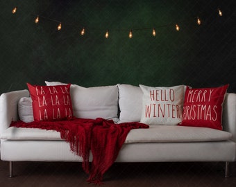 May include: A white couch with red throw pillows and a red blanket. The pillows have the text "Fa La La La La La La La Ja", "Hello Winter", and "Merry Christmas" on them. The couch is in front of a green wall with string lights.