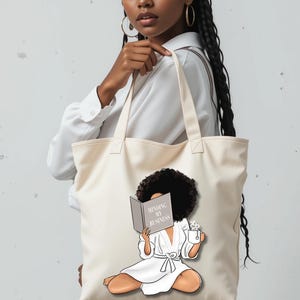 May include: A cream tote bag with an illustration of a woman reading a book with the text "Minding My Business." The woman is in a white robe and holding a cup. The bag is carried by a person.