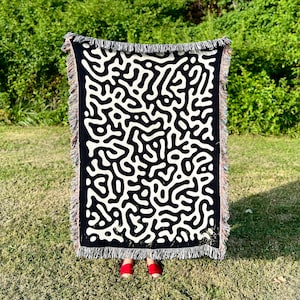Könnte beinhalten: Schwarz-weiße gewebte Wolldecke mit einem geometrischen Muster aus abstrakten Formen. Die Decke hat an allen Seiten Fransen.