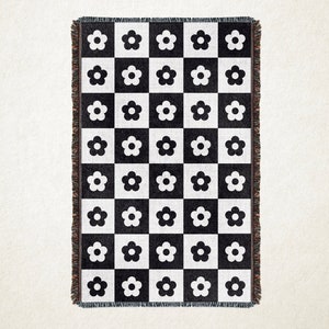 May include: A black and white woven throw blanket with a checkerboard pattern. Each square features a white flower. The blanket has a fringed edge. This decorative blanket is suitable for a bed or sofa.