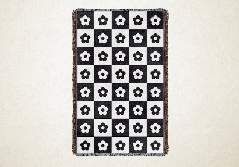 May include: A black and white woven throw blanket with a checkerboard pattern. Each square features a white flower. The blanket has a fringed edge. This decorative blanket is suitable for a bed or sofa.