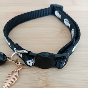 Peut inclure: Collier noir pour chat avec motif d'arêtes de poisson blanches. Boucle en plastique noir avec un visage de chat. Un charm en forme de poisson doré et une clochette noire sont attachés.