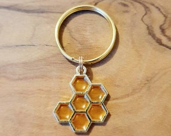 Puede incluir: Llavero dorado con un dije de panal de abeja. El dije es una representación detallada de un panal de abeja con un contorno dorado y un centro amarillo.
