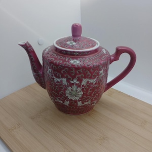 May include: A ceramic teapot featuring a deep magenta base colour with an intricate white and light grey swirling scrollwork pattern. The teapot includes a matching lid with a knob handle, a curved spout, and a sturdy handle, all decorated with floral motifs.